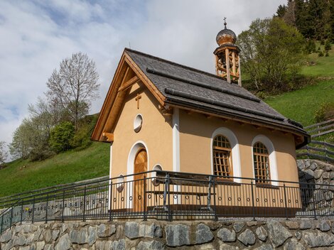 Eine kleine Kapelle auf einem sanften Hügel umgeben von Bäumen. Der Himmel ist teilweise bewölkt, und die Architektur ist schlicht und charmant. | © Bernhard Gruber – Photography