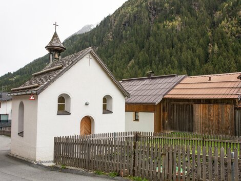 Eine kleine, weiß gestrichene Kirche mit einem spitzen Dach steht in einer ländlichen Umgebung. Im Hintergrund sind grüne Berge und traditionelle Holzhäuser zu sehen. | © Bernhard Gruber – Photography