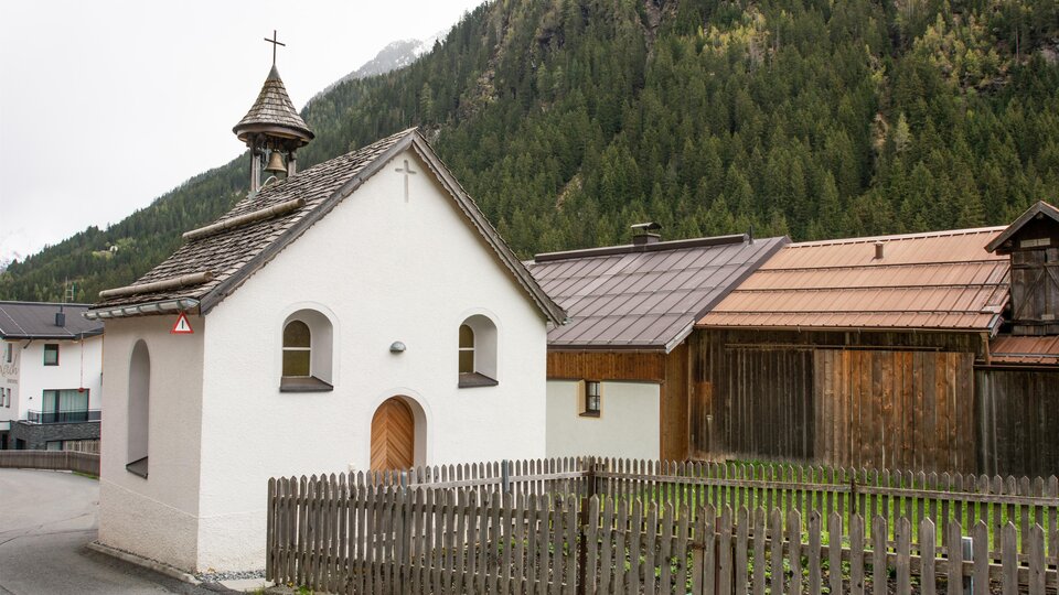 Eine kleine, weiß gestrichene Kirche mit einem spitzen Dach steht in einer ländlichen Umgebung. Im Hintergrund sind grüne Berge und traditionelle Holzhäuser zu sehen. | © Bernhard Gruber – Photography