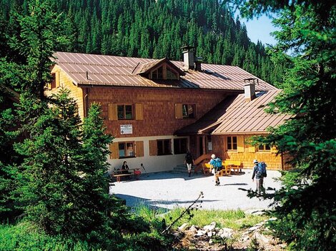 A mountain cabin surrounded by tall fir trees. In the foreground, hikers can be seen enjoying nature.