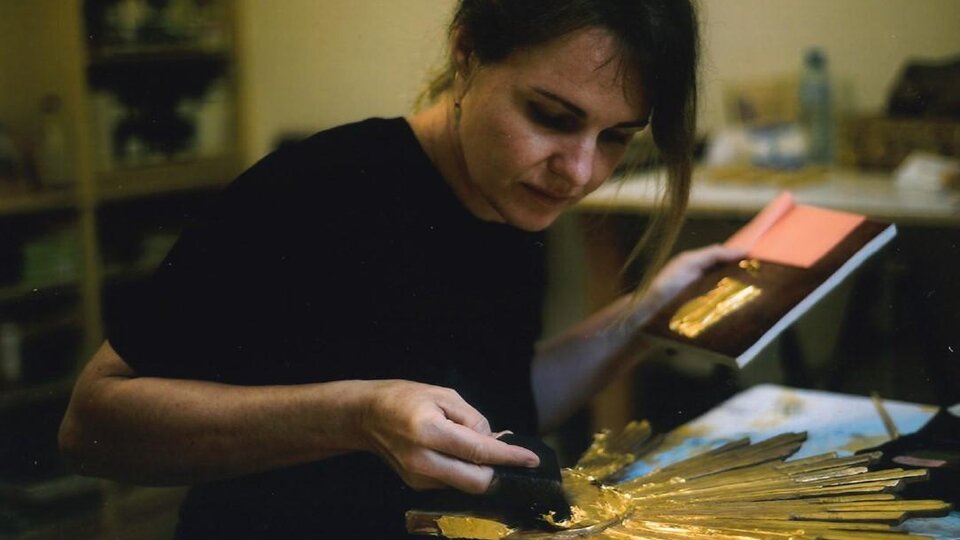 Eine Künstlerin arbeitet an einer goldfarbenen, strahlenförmigen Skulptur. Sie hält ein Buch in der Hand und konzentriert sich auf ihre kreative Tätigkeit.