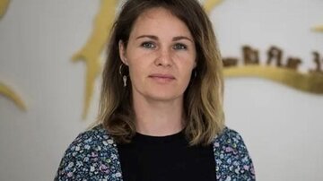 Eine Frau mit schulterlangen, lockigen Haaren blickt direkt in die Kamera. Im Hintergrund sind goldene dekorative Elemente sichtbar.