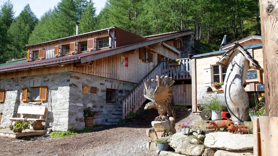 Ein gemütliches Holzhaus in den Bergen, umgeben von grünen Bäumen. Vor dem Haus steht eine dekorative Skulptur.