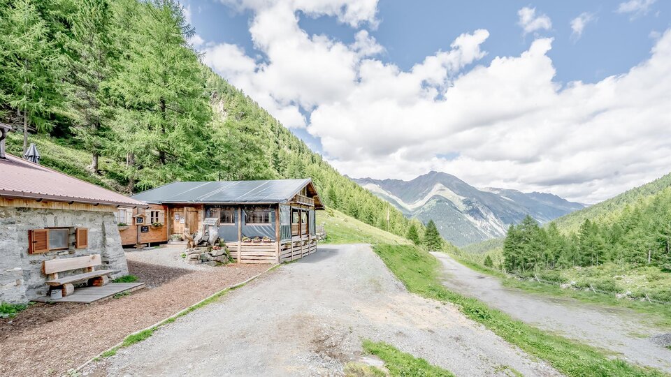 Eine malerische Alpenlandschaft mit zwei Hütten und grünen Bäumen. Die Berge im Hintergrund sind unter einem klaren Himmel sichtbar.
