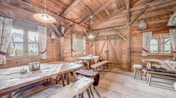 Eine gemütliche Holzstube mit einem großen Tisch und Bänken. Die Wände sind aus Holz, und durch die Fenster sieht man die Natur draußen.