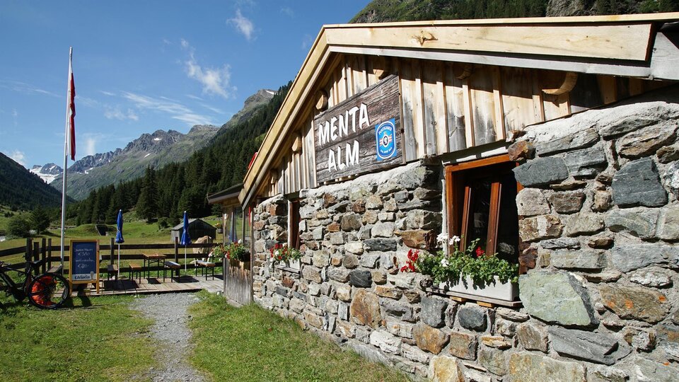 Eine gemütliche Almhütte aus Stein in einer malerischen Berglandschaft. Die Umgebung ist grün, mit Bergen im Hintergrund und blauer Himmel.