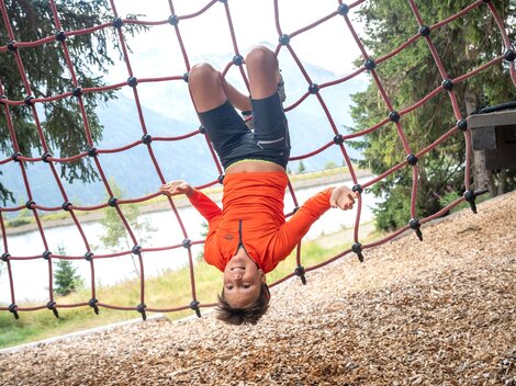 Ein Junge hängt kopfüber in einem roten Kletternetz und blickt fröhlich in die Kamera. Im Hintergrund sind Bäume, ein See und die verschwommene Berglandschaft zu sehen. Das Bild fängt einen spielerischen Moment voller Energie, Mut und Leichtigkeit im alpinen Outdoor-Erlebnis ein.