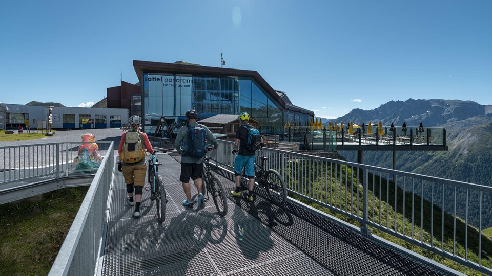 Drei Radfahrer stehen auf einer Aussichtsplattform mit Blick auf die Berge. Im Hintergrund ist ein modernes Gebäude zu sehen.