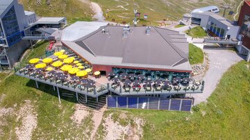 Ein modernes Restaurant auf einem Berg mit einer Terrasse und vielen Sonnenschirmen. Die Umgebung ist grün und bergig, ideal für einen entspannten Aufenthalt.