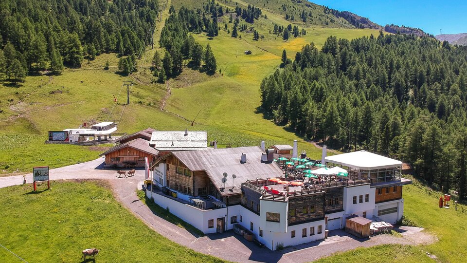 Ein gemütliches Bauernhaus inmitten einer grünen Berglandschaft. Im Hintergrund sind bewaldete Hänge und ein blauer Himmel zu sehen.