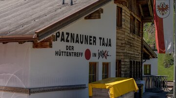 Ein traditionelles Almhütte-Schild mit der Aufschrift "Paznauner Taja - der HüttenTreff in Ischgl". Die Hütte ist aus Holz und umgeben von grünen Bergen.