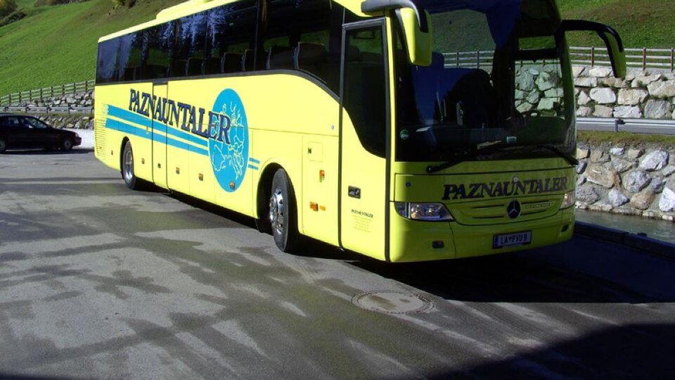 Ein gelber Reisebus steht an einem Parkplatz in einer bergigen Landschaft. Im Hintergrund sind grüne Hügel und einige Häuser zu sehen.