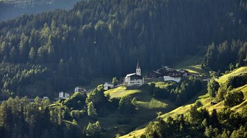Eine malerische Berglandschaft mit einem kleinen Dorf und einer Kirche. Umgeben von dichten Wäldern und grünen Wiesen.