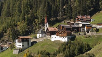 Ein malerisches Dorf in den Bergen, umgeben von grünen Wiesen und hohen Bäumen. Die Kirche mit dem charakteristischen Turm ist ein Blickfang in der Landschaft.