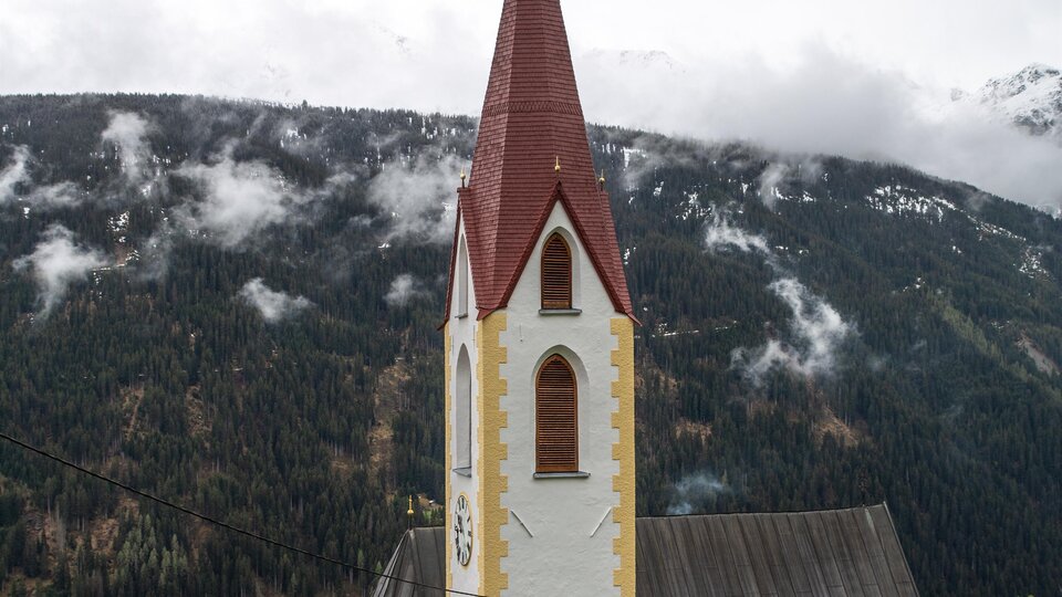 Eine Kirche mit einem roten Dach und einem hohen Kirchturm vor einer bergigen Landschaft. Die Umgebung ist von grünen Wäldern und sanften Hügeln geprägt. | © Bernhard Gruber – Photography