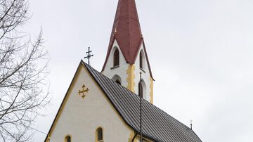 Eine schöne Kirche mit einem spitzen roten Turm und einem hellen Fassadenanstrich. Im Vordergrund sind einige Bäume und ein Zaun zu sehen. | © Bernhard Gruber – Photography