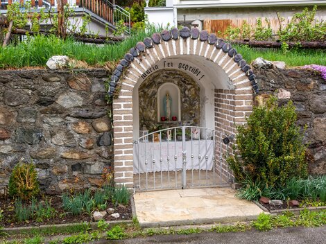 Ein kleiner Altarschrein in einer Steinmauer mit einem weißen Tor. Umgeben von grünen Pflanzen und Blumen. | © Bernhard Gruber – Photography