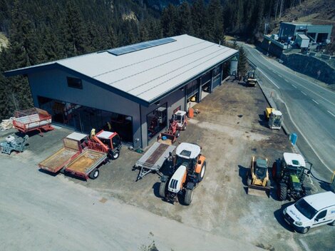 An agricultural workshop with several tractors and trailers on a property. The surroundings are surrounded by trees and mountains.