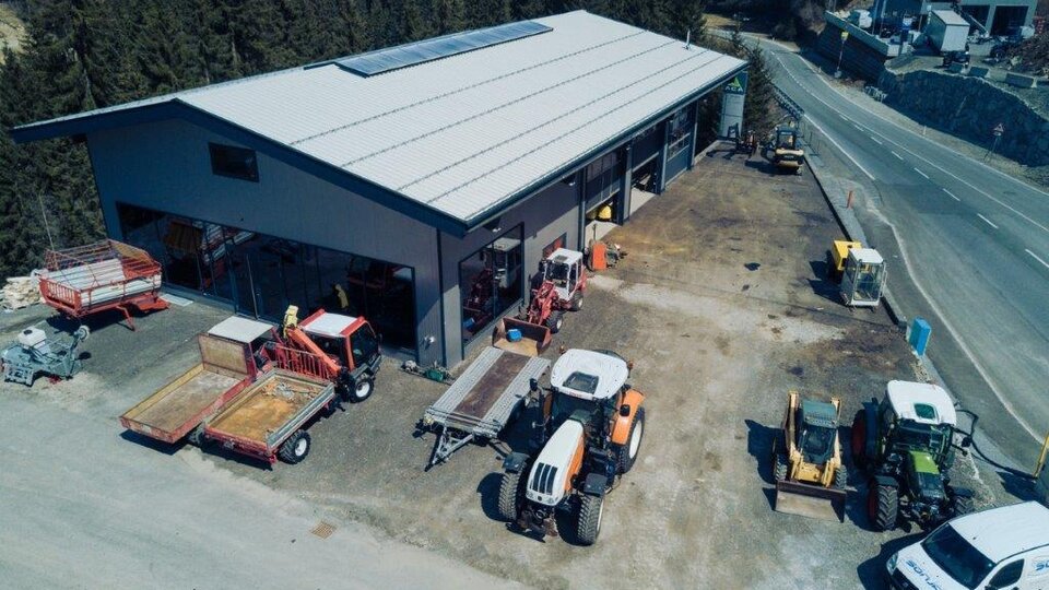 An agricultural workshop with several tractors and trailers on a property. The surroundings are surrounded by trees and mountains.