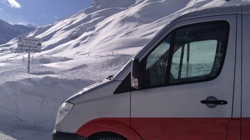 A van stands in front of a snowy landscape. In the background, snow-covered mountains and a road sign are visible.