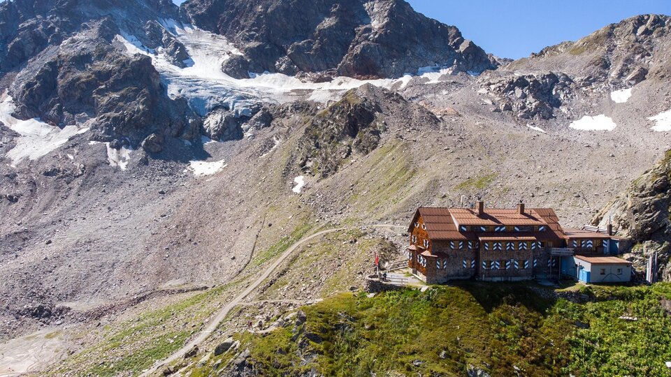 Eine Berghütte umgeben von beeindruckenden Bergen und Gletschern. Die Landschaft ist grün und bietet einen klaren Blick auf die umliegenden Gipfel.