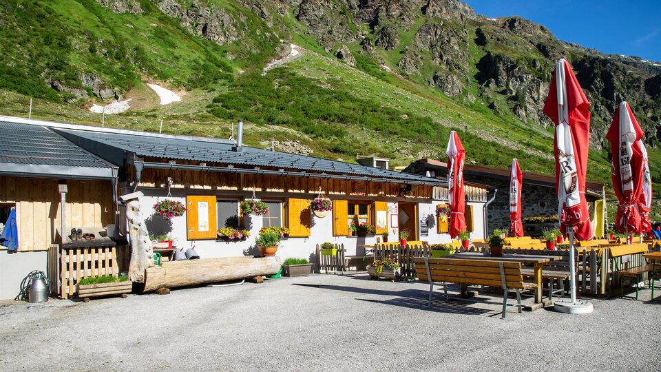 Eine gemütliche Almhütte mit bunten Blumen und roten Schirmen. Im Hintergrund sind grüne Berge zu sehen.