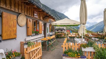 Eine gemütliche Berghütte mit Sonnenschirmen und Tischen im Freien. Im Hintergrund sind majestätische Berge und ein bewölkter Himmel zu sehen.