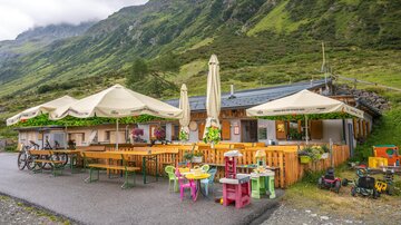 Eine gemütliche Berghütte mit Tischen und Sonnenschirmen im Freien. Die Umgebung ist grün und bergig, ideal für Ausflüge in die Natur.