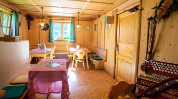 Ein gemütlicher Stubenbereich mit Holzwänden und einem Tisch mit karierter Tischdecke. Die Fenster sind mit blauen Vorhängen geschmückt und bieten einen Blick ins Freie.