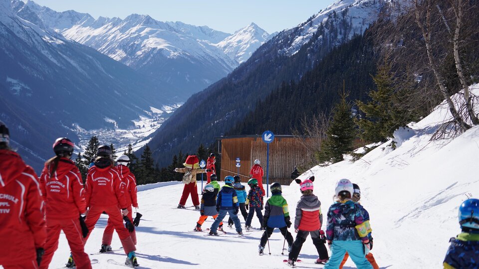 Eine Gruppe von Kindern und Skilehrern auf einer Skipiste in den Bergen. Die Umgebung ist verschneit und es ist sonnig.