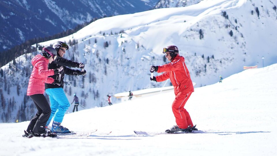 Eine Skischule in den Bergen mit drei Personen, die Skifahren lernen. Die Umgebung ist schneebedeckt und die Berge sind beeindruckend.