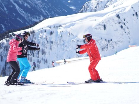 Three people in ski lessons on a snowy slope. Impressive mountains can be seen in the background.