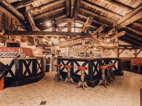 Eine gemütliche Bar in einem rustikalen Ambiente mit Holzdecken und -balken. Rote Barhocker umgeben die zentrale Theke.