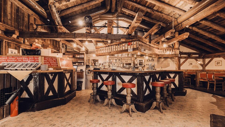 Eine gemütliche Bar in einem rustikalen Ambiente mit Holzdecken und -balken. Rote Barhocker umgeben die zentrale Theke.