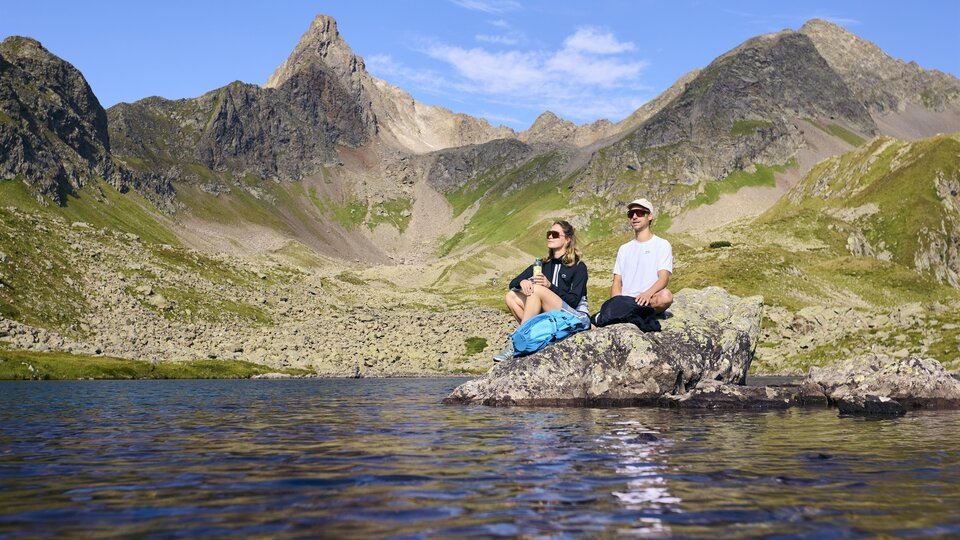 Zwei Personen sitzen auf einem Felsen am Ufer eines Sees in den Bergen. Die Landschaft ist majestätisch mit hohen Gipfeln und klarem Himmel.