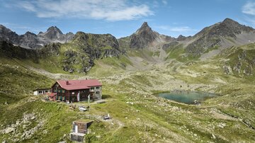 Eine malerische Berglandschaft mit einer Hütte und einem kleinen See. Umgeben von hohen Bergen und grünem Gras.