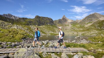 Zwei Wanderer gehen über einen Holzsteg in einer malerischen Berglandschaft. Im Hintergrund sind grüne Wiesen und hohe Berge zu sehen.