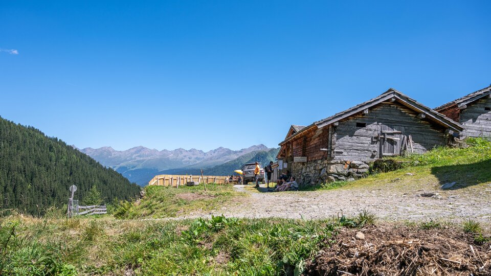 Ein malerisches Bergdorf mit traditionellen Holzgebäuden und einer wunderschönen Aussicht auf die Berge. Der strahlend blaue Himmel und die grüne Wiese verleihen der Szene eine serene Atmosphäre.