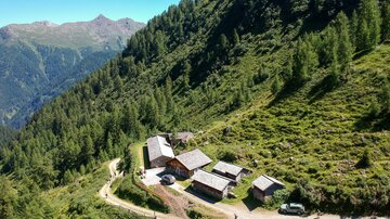 Eine idyllische Berghütte umgeben von grünen Wiesen und Bäumen. Der Weg führt sanft durch die Landschaft in die Berge.