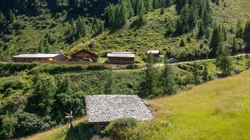 Eine malerische Berglandschaft mit grünen Wiesen und Wäldern. Im Vordergrund ist eine Hütte zu sehen, während im Hintergrund weitere Gebäude stehen.