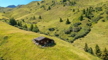 Eine malerische Hütte steht auf einer sanften Wiese in den Bergen. Grüne Hügel und klare blaue Himmel umgeben die Szene.