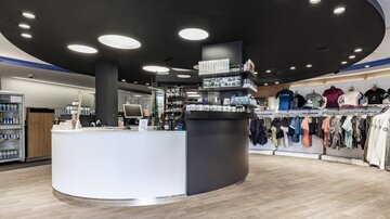 Ein moderner Verkaufsraum mit einem runden Empfangstresen und einer Kleidungsauslage. Die Wände sind hell gestaltet, und die Beleuchtung ist freundlich und einladend.