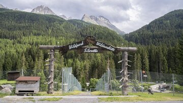 An entrance to the wildlife park with a large wooden sign. In the background, green forests and mountains are visible.
