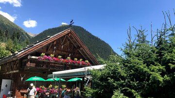 A charming wooden house with colorful flower boxes. In the foreground, people sit under sun umbrellas in a picturesque mountain landscape.