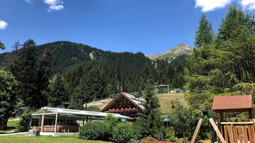 A picturesque mountain village with a traditional building and lush trees. In the background, there are green hills and a blue sky.