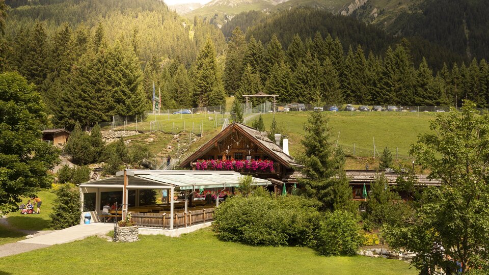 A cozy mountain restaurant surrounded by green meadows and tall trees. In the background, majestic mountains and clear skies can be seen.
