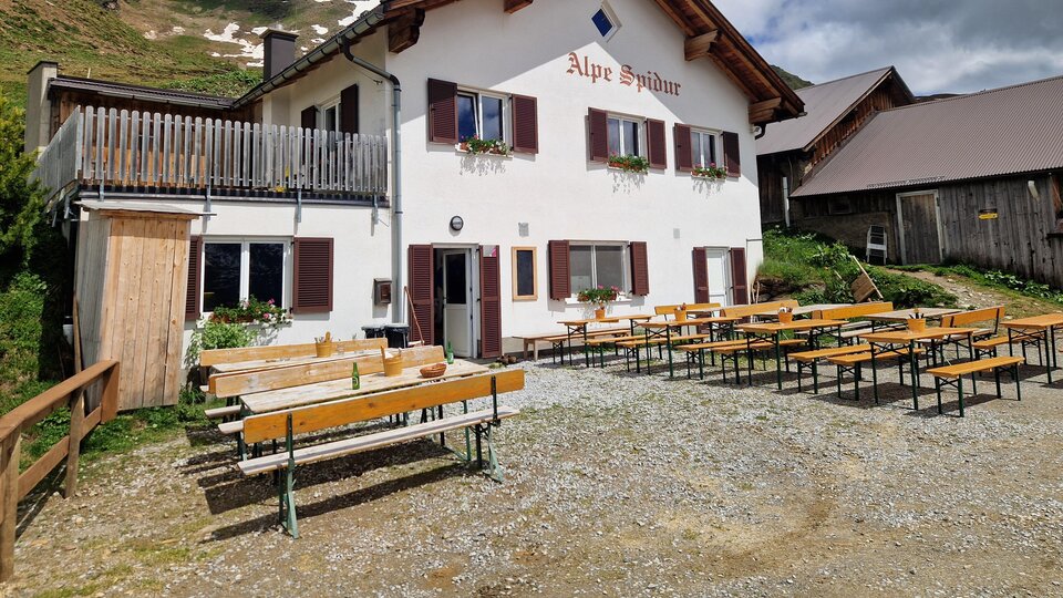 Ein gemütliches Alpenhaus mit dem Namen "Alpe Spina". Draußen stehen viele Bänke und Tische in einer malerischen Berglandschaft.