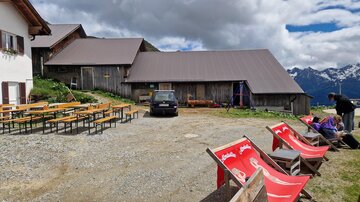 Ein rustikales Berglokal mit einer großen Terrasse. Im Vordergrund stehen Liegestühle und Holztische, während im Hintergrund die Berge und Wolken sichtbar sind.