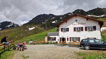 Ein gemütliches Alpenhaus umgeben von hohen Bergen. Auf dem Gelände sind einige Sitzgelegenheiten und Autos zu sehen.