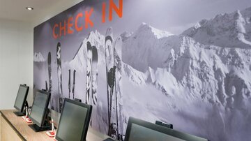 Ein moderner Check-in-Bereich mit mehreren Computern. Im Hintergrund sind Skier vor einer schneebedeckten Berglandschaft abgebildet.